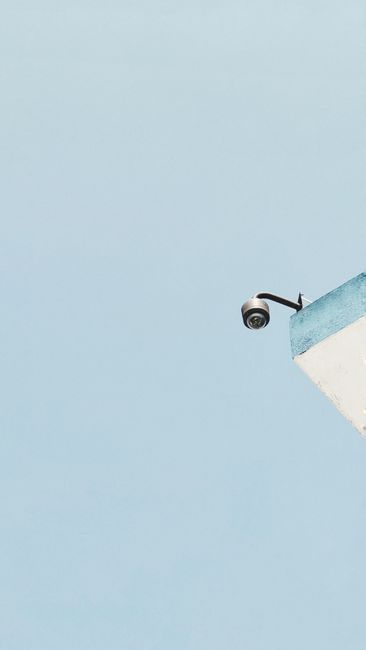 A low-angle view of a security camera on a blue and white building corner against a blue sky.